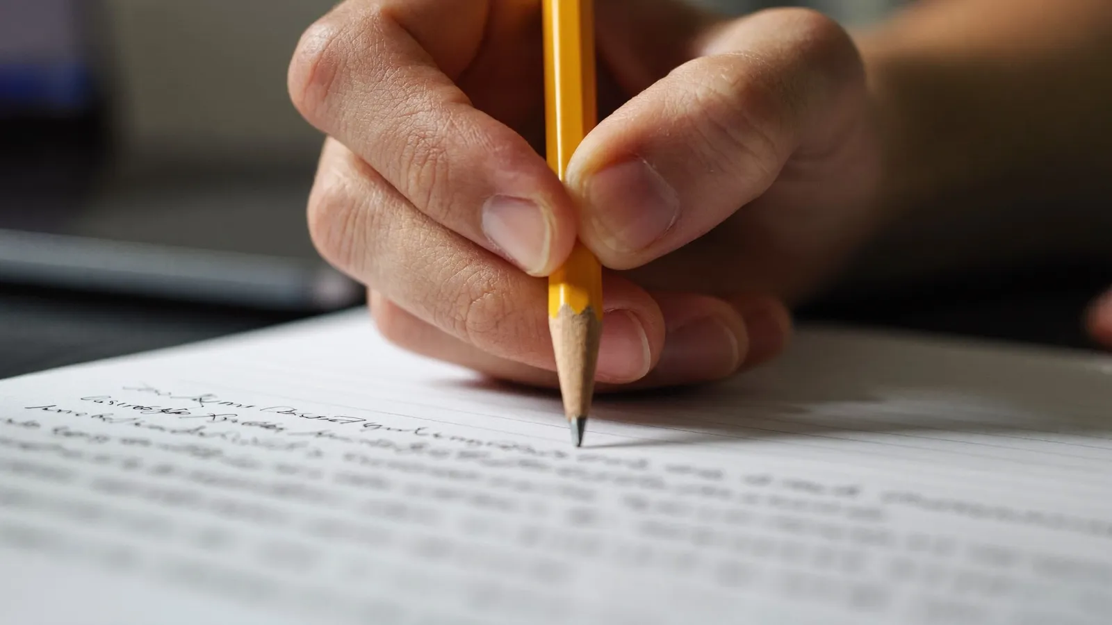 Close-up of a hand writing in pencil on a lined notebook page, with a laptop out of focus behind it