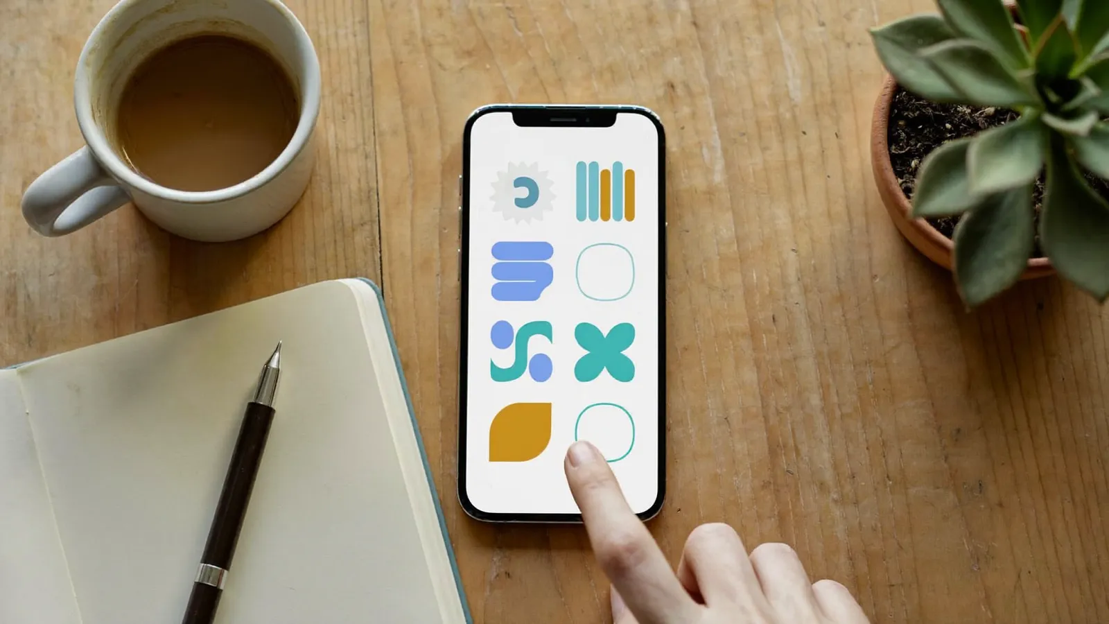 Overhead view of a desk with a hand about to tap a smartphone showing a grid of colorful abstract app icons, next to a coffee mug, an open notebook, and a small plant
