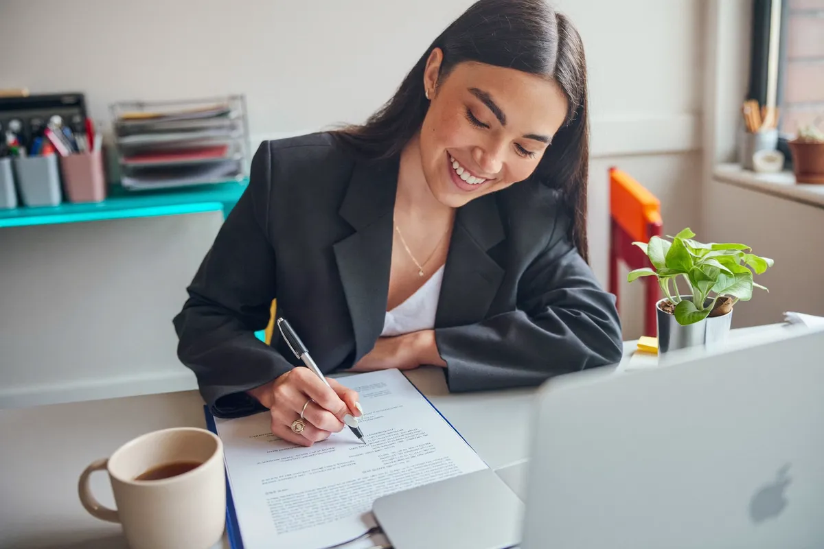 A smiling office worker marking small edits on a printed draft with a pen, a coffee mug and a plant on the desk