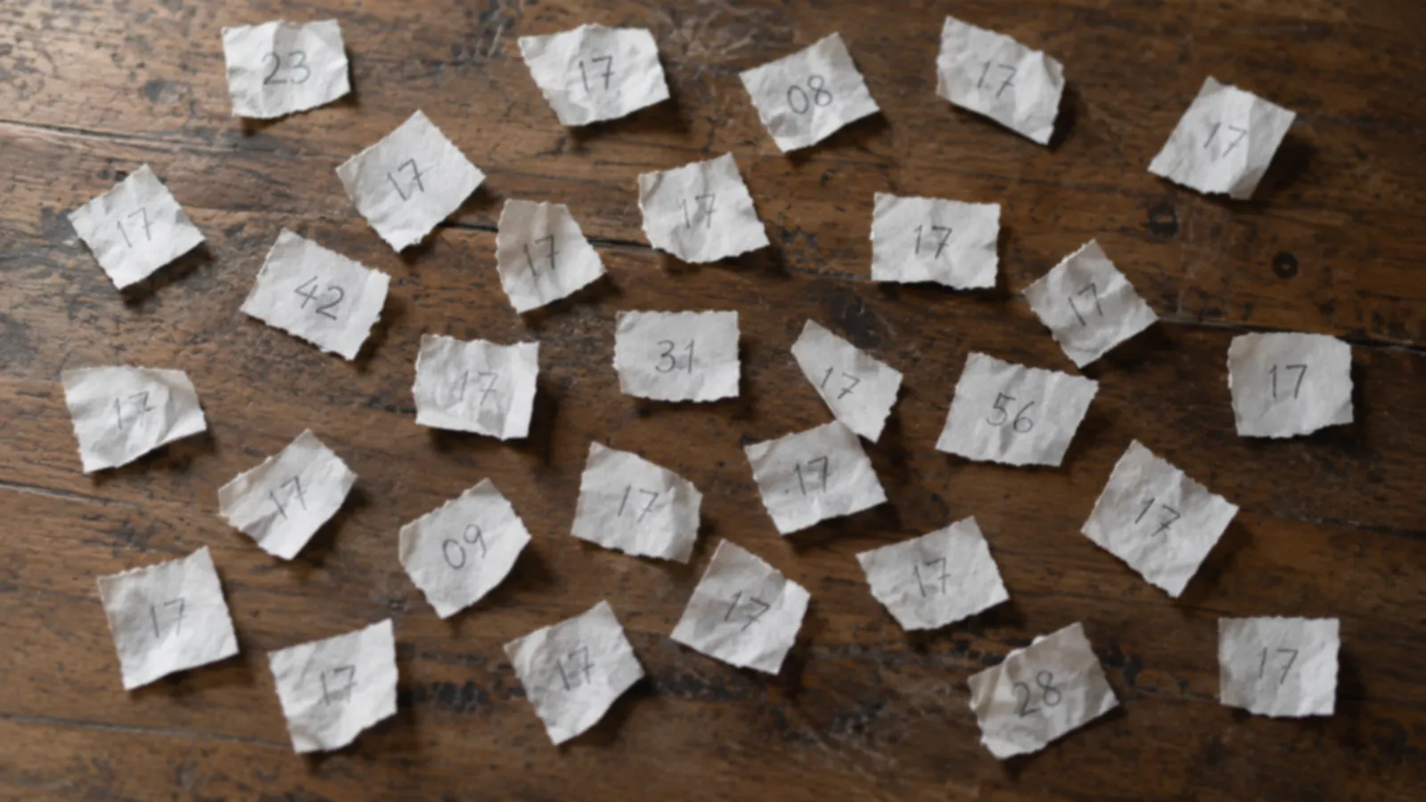 An overhead photograph of a wooden desk covered in small torn paper slips, each with a number handwritten in pencil, and most of them reading 17