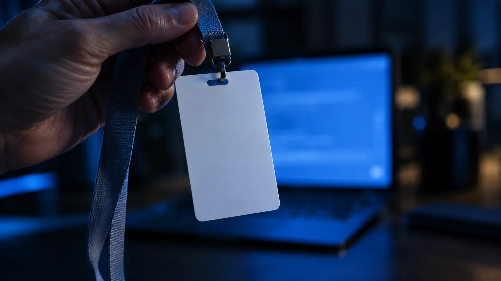A hand holding up a completely blank white ID badge on a lanyard, in front of a blue-lit laptop screen