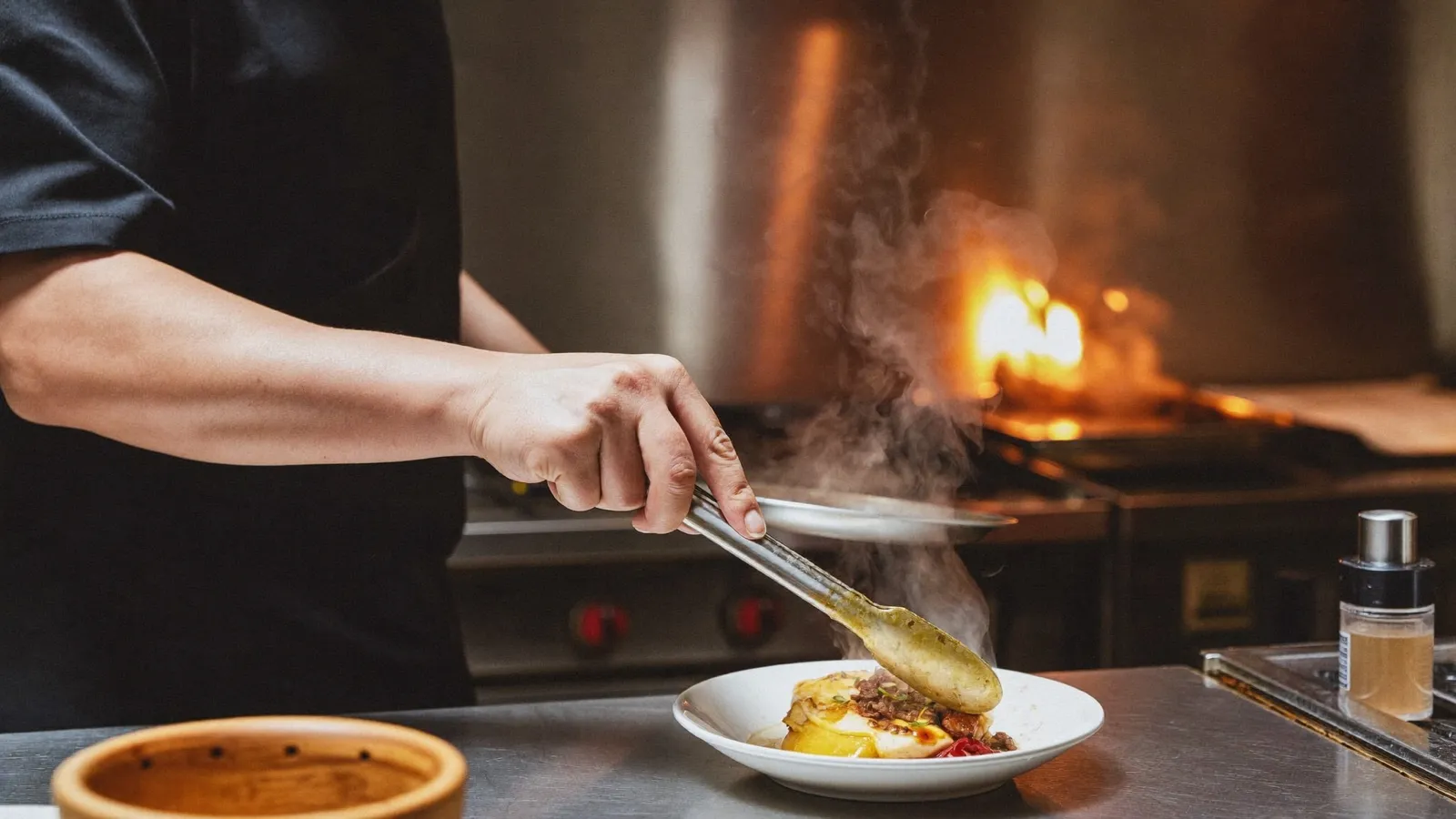 A cook's hands plating a dish with tongs over a hot pass in a restaurant kitchen, steam rising and a burner flame behind