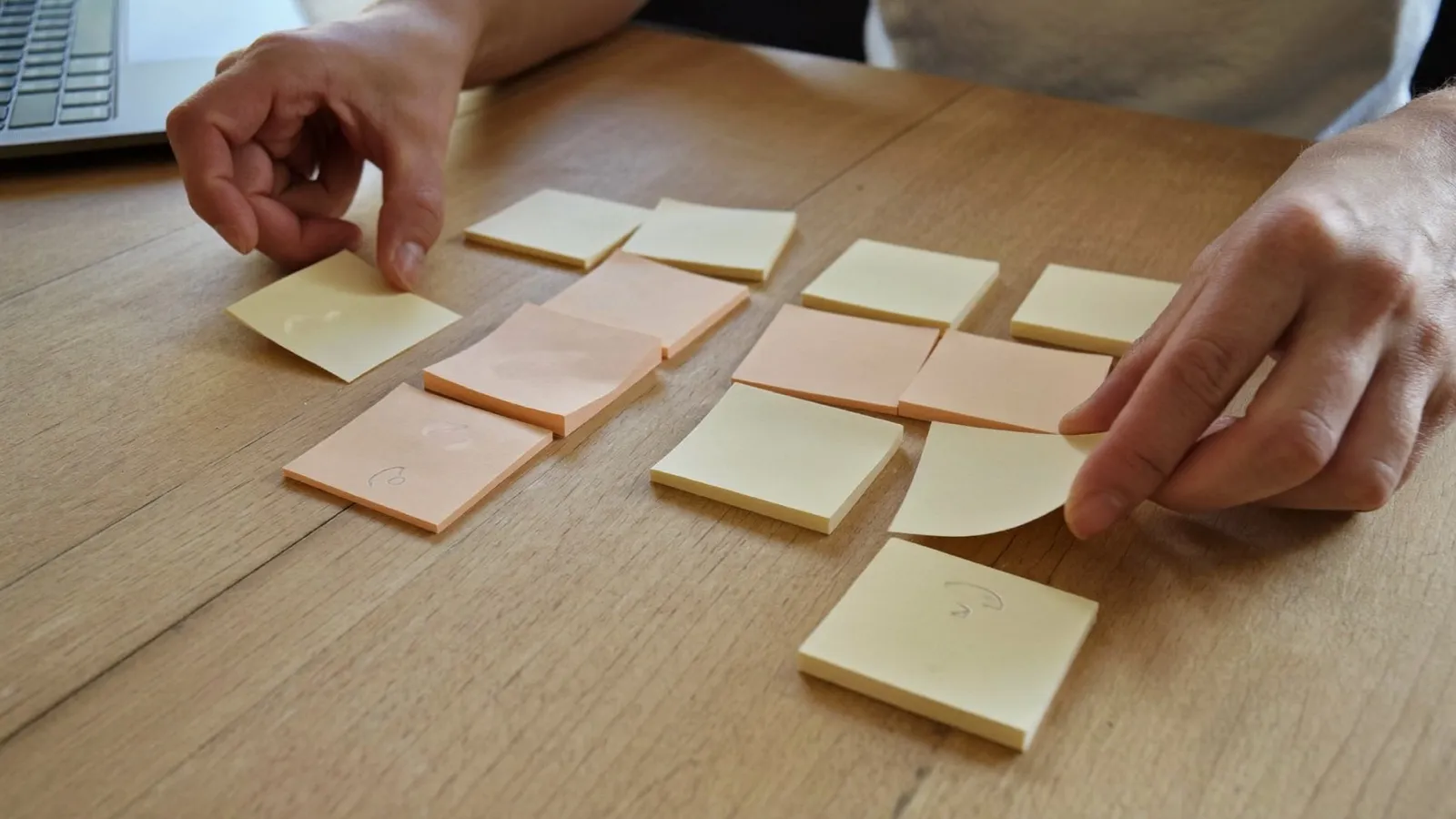 Two hands sorting blank sticky notes into separate groups on a wooden desk beside a laptop